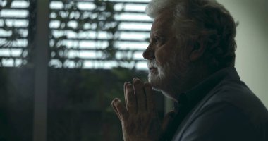 Derin yansıtıcı dualarda el ele tutuşan yaşlı bir adam, yumuşak ışık ve kasvetli atmosferiyle pencerenin yanında sessizce duruyor.