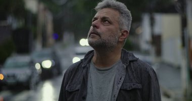 Orta yaşlı bir adam yağmurun altında duruyor. Islak giysiler ve kasvetli bir ifade, bulanık arabalar ve karamsar sokak atmosferiyle çevrili.