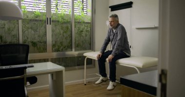 Muayenehanede muayene masasında tek başına oturan orta yaşlı bir adam. Muayeneden sonra düşünceli ve sıkıntılı görünüyor. Klinik kaplıcada sakin bir yansıma anı.