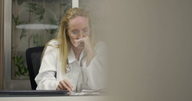 Kadın doktor masasında oturuyor. Yorgunluk ve duygusal yorgunluk gösteriyor. Gözler kapalı ve elleri klinikte iş sırasında yüzün üzerinde.