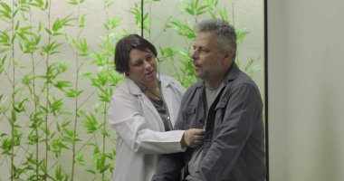Doktor, orta yaşlı bir erkek hastanın göğsünde steteskop kullanıyor. Klinik çevrede, yapraklı arka planı olan tıbbi muayene sırasında.