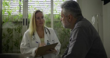 Muayene odasında orta yaşlı bir hastayla yapılan görüşmeler sırasında doktor tıbbi notları inceliyor. Hasta öne eğiliyor. Endişe ve dikkatle dinliyor.