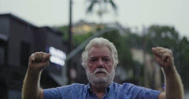 Yaşlı bir adam yağmurda kolları hafifçe havada duruyor, kameraya geniş gözlerle bakıyor ve şok, inançsızlık ve duygusal hassasiyet ifadesi sergiliyor.