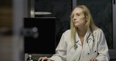 Kadın doktor bilgisayar ekranına odaklanmış, geceleri klinikte klavyeye yazı yazıyor, reçeteli şişeler ve hasta belgelerine bakılıyor.