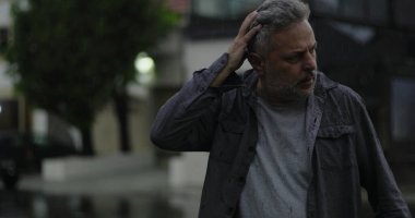 Orta yaşlı bir adam, yağmurun altında, elleri başının üstünde, gözleri açık, hayal kırıklığı ve zihinsel yorgunluk içinde.