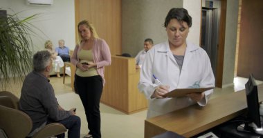 Hastalar arka planda beklerken ve çalışanlar yoğun tıbbi lobide ziyaretçilerle uğraşırken klinik resepsiyonunda not alan kadın doktor.