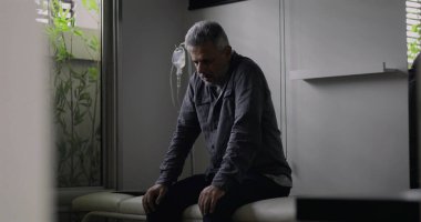 Klinik odasında tek başına oturan, arkasında serum olan, sessizce aşağıya bakan, tıbbi konsültasyondan ya da teşhisten sonra duygusal olarak ağırlığını koyan bir adam.