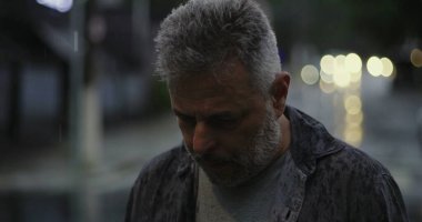 Orta yaşlı bir adam yağmurda aşağıya bakıyor, gözleri derin duygusal bir yansımayla kapalı, sırılsıklam gömlek ve bulanık şehir ışıkları altında.