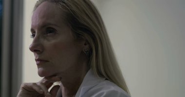 Yan profildeki kadın doktor, elleri çenesinin altında, gözleri klinik bir çevrenin içinde derin bir yansıma anında ileri bakıyor.