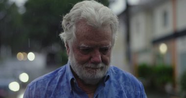 Yaşlı bir adam yağmurun altında üzgün gözlerle ve duygusal ifadeyle, dudakları sımsıkı ve kasvetli bir şekilde duruyor.