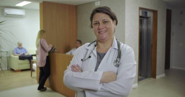 Klinik koridorunda kolları çapraz gülümseyen, steteskop ve laboratuvar önlüğü giyen, resepsiyondaki ve arka gruptaki hastalar tarafından görülebilen kendinden emin bir kadın doktor.