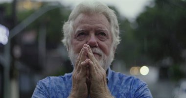 Yüzüne yapışmış elleri tutan yaşlı bir adam, yağmurda parlayan gözler, duygu, umut, ya da ruhsal bağlantı anında sessiz dua ile dolu.