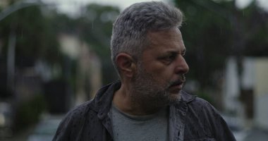 Orta yaşlı bir adam endişeli bir ifadeyle, gergin duruşu ve şehir sokaklarında duygusal ağırlığıyla yan yana bakıyor.