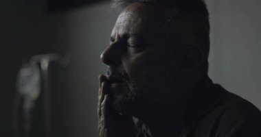 Klinikte gözleri kapalı oturan orta yaşlı bir adamın yakın plan çekimi, tıbbi belirsizlik anında sessizce ağrıyı ve yorgunluğu işliyor.