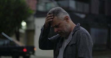 Orta yaşlı bir adam, yağmurun altında, elleri başının üstünde, gözleri açık, hayal kırıklığı ve zihinsel yorgunluk içinde.