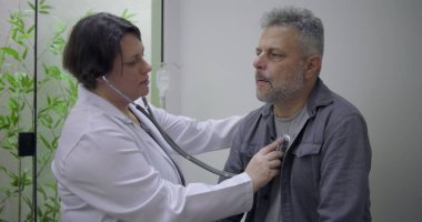 Steteskop kullanan kadın doktor klinik ortamdaki rutin sağlık kontrolleri sırasında orta yaşlı erkek hastaların kalp atışlarını inceliyor.
