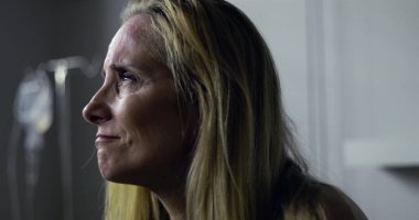 Klinikteki kadının yoğun, duygusal bir ifadeyle yan tarafa baktığı yakın plan. Arkaplanda tıbbi tedavi veya iyileşme öneren serum var.