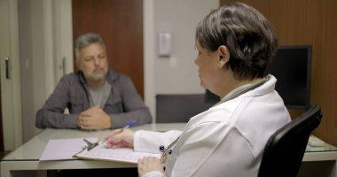Doktor, tıp bürosunda kağıt ve cam masadaki belgelerle baş başa görüşmede orta yaşlı bir hastaya hitap ediyor.