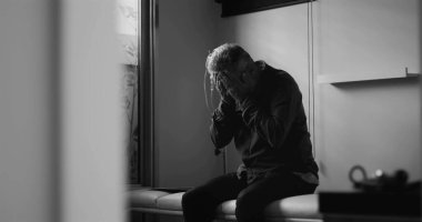 Klinikte başı dertteyken oturan orta yaşlı bir adam, dramatik siyah beyaz görüntü, tıbbi çevrede duygusal acı ya da zihinsel yük taşıyor.