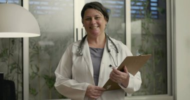 Stetoskop ve not panosuyla gülümseyen kadın doktor klinik ofisinde rahat duruşu ve modern dekoruyla duruyor.