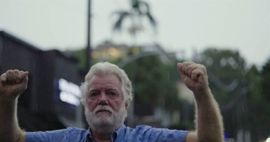 Yaşlı bir adam yağmurda kolları hafifçe havada duruyor, kameraya geniş gözlerle bakıyor ve şok, inançsızlık ve duygusal hassasiyet ifadesi sergiliyor.