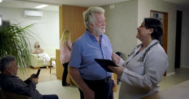 Kadın doktoru dinlerken gülümseyen kıdemli adam klinik lobisindeki dijital tablet hakkında bilgi veriyor, resepsiyon masasının yanındaki günlük sağlık danışmanlığı