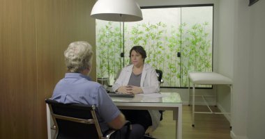 Klinik ofis ortamındaki yaşlı erkek hastaya teşhisi açıklayan doktor güven, iletişim ve profesyonel sağlık aracına odaklanmış ciddi tıbbi danışmanlık