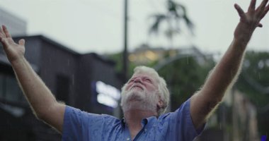 Kolları geniş ve yüzü göğe yükselmiş yaşlı bir adam, ruhsal rahatlama, minnettarlık veya duygusal teslimiyet ifadesiyle yağmuru kucaklıyor.