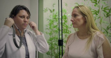 Orta yaşlı bir kadının göğsünü steteskopla inceleyen kadın hekim modern klinikte bitki duvarlı arka gruplaşmayla sağlık muayenesi sırasında