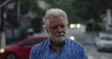 Yaşlı bir adam yağmurun altında üzgün gözlerle ve duygusal ifadeyle, dudakları sımsıkı ve kasvetli bir şekilde duruyor.