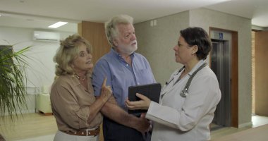 Üst düzey çift, klinik koridorundaki olumlu sağlık gelişmelerinden sonra kadın doktorla neşe içinde gülümsüyor. Rahatlama ve mutluluk dile getiriyorlar.