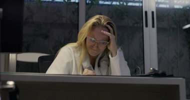 Yorgun kadın doktor, masa başında oturuyor. Uzun bir vardiyadan sonra bunalmış ve stresli. İş yerinde bitkinlik ve yorgunluğun duygusal ifadesi.