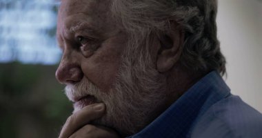 Düşünceli bir anda yaşlı bir adam, elleri çenesinde, düşünceli bir ifadeyle dışarı bakıyor, evin içinde hafif ışıklı pencere ve bulanık bir arka plan.