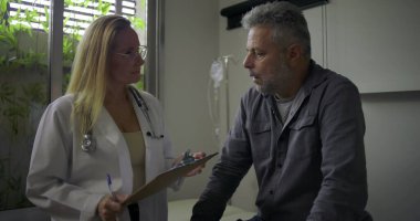 Kadın doktor klinik muayenesinde orta yaşlı erkek hastaya nazikçe kötü haber veriyor.