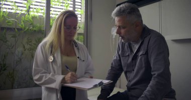 Parlak tıp ofisinde muayene masasında oturan orta yaşlı bir hastaya danışmanlık yaparken elinde dosya tutan kadın doktor.