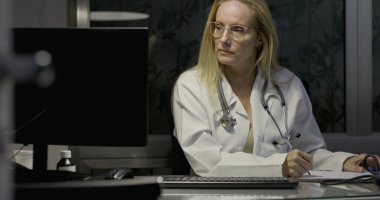 Kadın doktor bilgisayar ekranına odaklanmış, geceleri klinikte klavyeye yazı yazıyor, reçeteli şişeler ve hasta belgelerine bakılıyor.