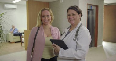 Gülümseyen doktor ve hasta klinik koridorunda yan yana duruyor, dijital tableti tutuyor ve parlak sağlık ortamında kameraya bakıyor.
