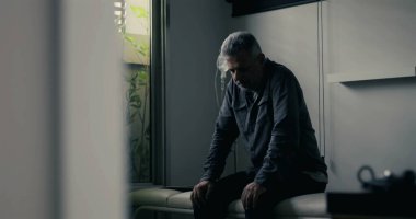 Sıkıntılı adam klinikte yalnız başına oturuyor, ağır duruşuyla öne eğiliyor, bastırılmış ışıklandırma yalnızlığı ve duygusal çöküntüyü vurguluyor.