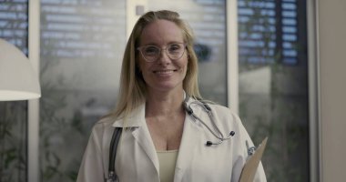 Elinde klasör ve steteskopla gülümseyen kadın doktor, modern klinikte iç mekan bitkileri ve büyük pencere paneli olan kameraya bakıyor.