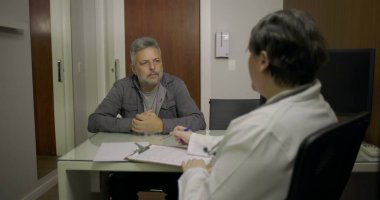 Orta yaşlı bir hasta klinikteki danışma sırasında doktorunu dikkatle dinliyor. Elinde not defteri ve masasında notlar var.