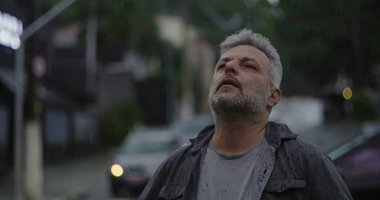 Orta yaşlı bir adam yağmurun altında duruyor. Islak giysiler ve kasvetli bir ifade. Etrafı bulanık arabalar ve kasvetli sokak atmosferiyle çevrili.