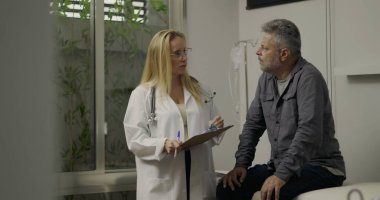 Klinikteki tıbbi randevu sırasında doktorla konuşan orta yaşlı erkek hasta, arka planda pano ve IV ekipmanıyla sağlık sorunlarını tartışıyor.