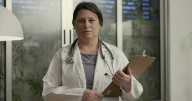 Modern sağlık hizmetleri bürosunda pano ve steteskopla çalışan profesyonel kadın doktorların ifadesiyle doğrudan kameraya bakması.