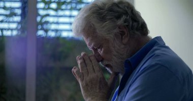 Pencerenin yanında duran mavi gömlekli yaşlı adam, gözleri kapalı ve elleri derin duygusal dualarla ya da hüzünlü yansımalarla birleşmiş.