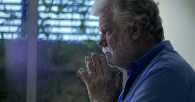 Yüzü nazik bir ifadeyle yukarı bakan yaşlı bir adam dua veya yansıma, elleri yavaşça birbirine yapışmış pencerenin yanında duruyor