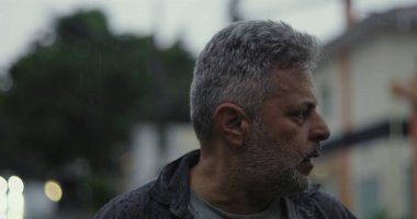 Orta yaşlı bir adam endişeli bir ifadeyle, gergin duruşu ve şehir sokaklarında duygusal ağırlığıyla yan yana bakıyor.