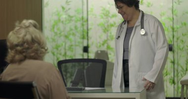 Steteskoplu gülümseyen doktor profesyonel klinikteki tıbbi danışmanlık sırasında yaşlı kadın hastayla samimi bir şekilde temas kuruyor.