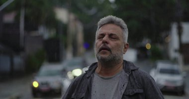 Orta yaşlı bir adam yağmurda dikiliyor, başı arkaya eğik ve gözleri kapalı, rahatlama, üzüntü veya duygusal katarsi ifade ediyor.