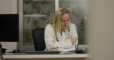 Masasında oturan kadın doktor iş saatinde klinikte bitkinlik ve duygusal yorgunluk belirtileri gösteriyor.