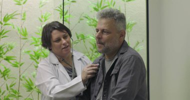 Doktor, klinik ortamda yapraklı arka grubuyla tıbbi muayene sırasında orta yaşlı bir erkek hastanın göğsünde steteskop kullanıyor.
